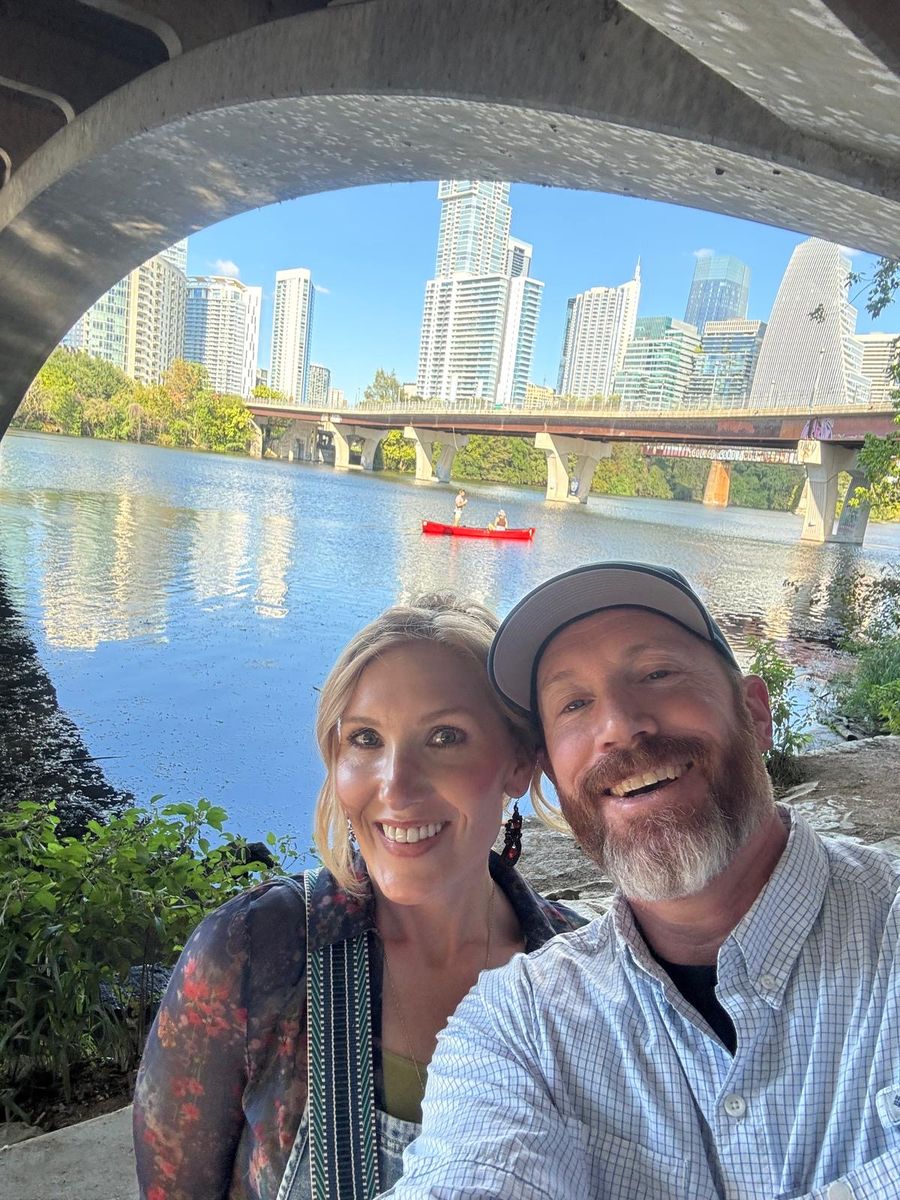 Tabitha and Kevin Lovell under a bridge arch on Lady Bird Lake, with the Austin skyline and a red canoe on the water behind them